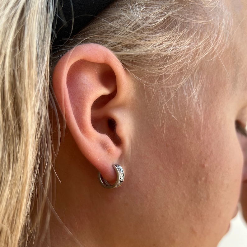 May include: A close-up of a person's ear with a small silver hoop earring with a decorative design. The earring is worn in the lower lobe of the ear.