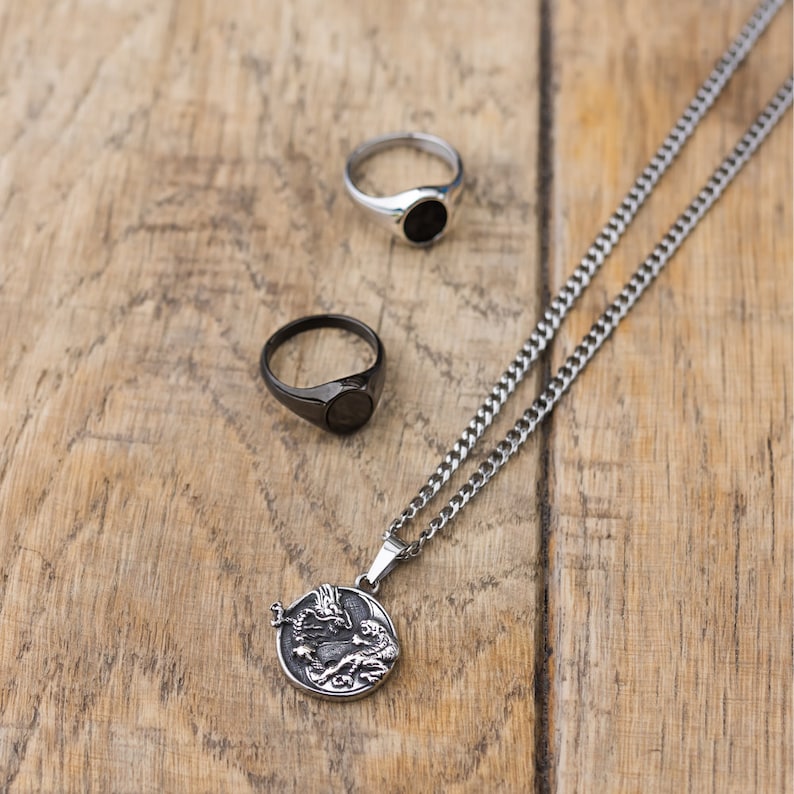 May include: A silver chain necklace with a black dragon pendant. The pendant is round and features a detailed design. The necklace is also paired with two black signet rings.