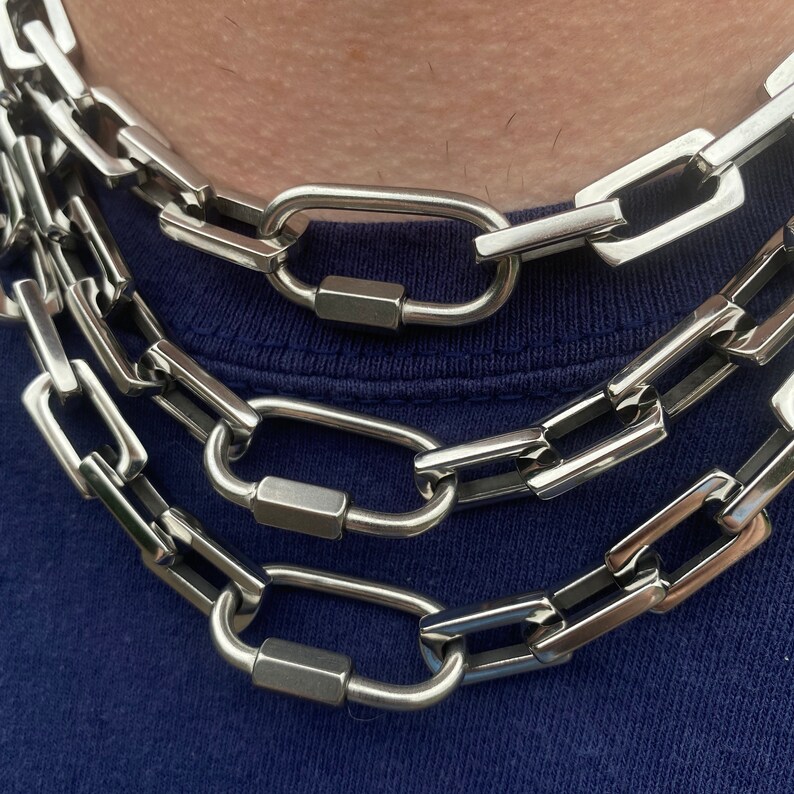 May include: Close-up of three silver-colored chain necklaces with large, rectangular links and oval-shaped clasps. The necklaces are layered and rest against a blue fabric background. The chains have a shiny, metallic finish.