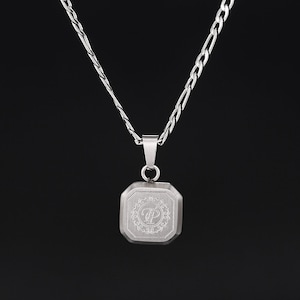 May include: A silver-colored necklace with a square pendant. The pendant has an engraved design with the letters "TP" inside a decorative border. The necklace chain is a combination of a curb and figaro style.