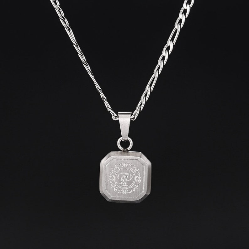 May include: A silver-colored necklace with a square pendant. The pendant has an engraved design with the letters "TP" inside a decorative border. The necklace chain is a combination of a curb and figaro style.