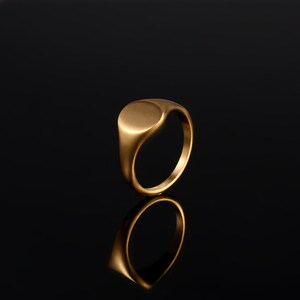 May include: A gold signet ring with a smooth, oval top. The ring is on a black background.