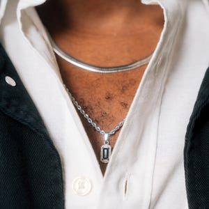 Könnte beinhalten: Eine silberne Halskette mit einem rechteckigen Anhänger mit einem dunklen Stein und kleinen umliegenden Steinen. Der Anhänger hängt an einer silbernen Kette. Eine zweite silberne Kette ist ebenfalls sichtbar. Die Halsketten werden über einem weißen Hemd getragen.
