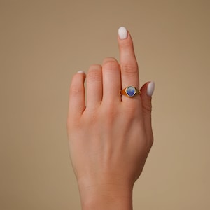 Op de afbeelding: Een gouden ring met een blauwe ovale edelsteen. De ring zit aan een vinger.