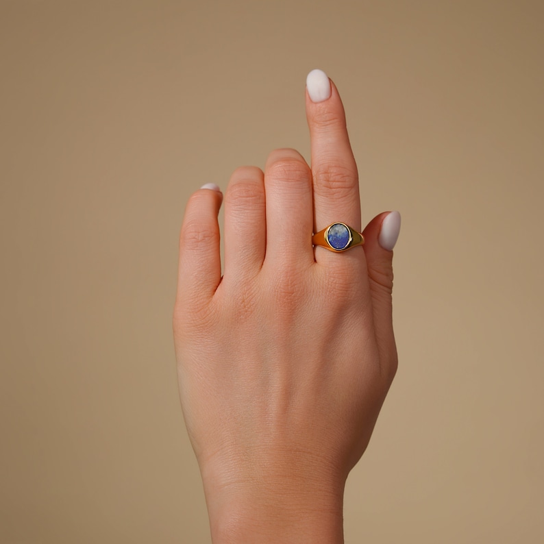 Op de afbeelding: Een gouden ring met een blauwe ovale edelsteen. De ring zit aan een vinger.