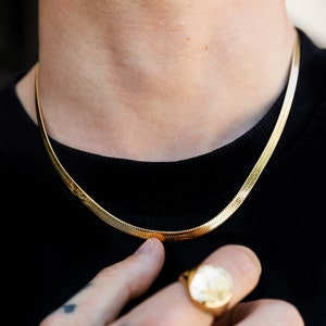 May include: A gold chain necklace with a snake chain design. The chain is worn by a person with a black shirt.
