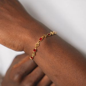 May include: Gold-toned bracelet featuring rectangular red gemstones and star-shaped accents. The bracelet is displayed on a wrist, showcasing its delicate design and color contrast. The jewelry piece is a stylish accessory.