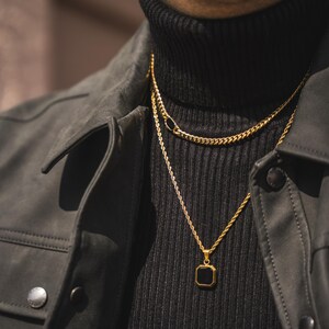 May include: A close-up of two gold necklaces. The top necklace is a curb chain, and the bottom necklace has a square black pendant. The necklaces are layered over a black turtleneck and a dark gray jacket.
