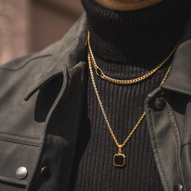 May include: A close-up of two gold necklaces. The top necklace is a curb chain, and the bottom necklace has a square black pendant. The necklaces are layered over a black turtleneck and a dark gray jacket.