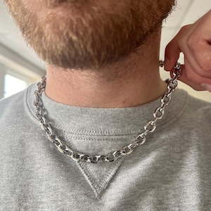 Könnte beinhalten: Eine silberne Kettenkette im Curb-Link-Stil. Die Kette ist dick und hat eine glänzende Oberfläche.