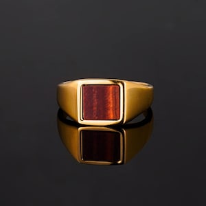 Könnte beinhalten: Ein goldfarbener Ring mit einem rechteckigen, rotbraunen Stein in der Mitte. Der Ring wird auf einer reflektierenden schwarzen Oberfläche präsentiert, wodurch ein Spiegeleffekt entsteht. Das Design des Rings ist schlicht und elegant.