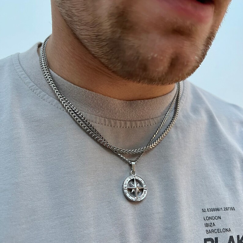 May include: A silver-colored necklace with a compass pendant. The pendant features a detailed compass design with a star in the center. The necklace is layered with a thicker chain and a thinner chain. The person is wearing a gray t-shirt.