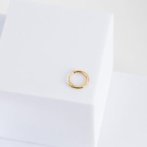 Puede incluir: Un solo pendiente de aro de color dorado. El pendiente es circular y tiene un dise&ntilde;o simple y minimalista. El pendiente est&aacute; sobre un fondo blanco.