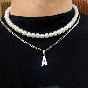 May include: A silver chain necklace with a silver pendant in the shape of the letter 'A'. The necklace is layered over a pearl necklace.
