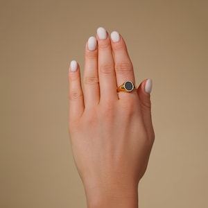 Op de afbeelding: Een goudkleurige zegelring met een zwarte ovale steen wordt op een vinger gedragen. De ring heeft een gladde, gepolijste afwerking. De achtergrond is effen en neutraal.