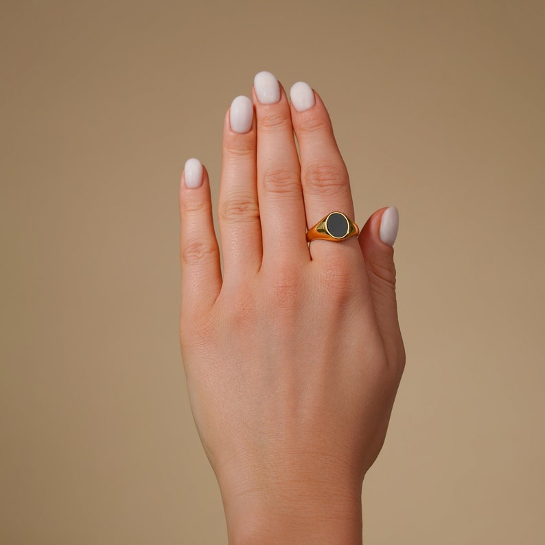 Op de afbeelding: Een goudkleurige zegelring met een zwarte ovale steen wordt op een vinger gedragen. De ring heeft een gladde, gepolijste afwerking. De achtergrond is effen en neutraal.