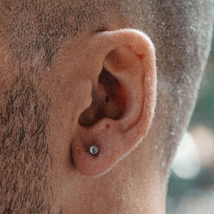 Puede incluir: Un pequeño pendiente de botón circular de tono plateado está colocado en el lóbulo inferior de una oreja. El pendiente presenta una superficie metálica lisa y pulida que refleja la luz, creando un brillo sutil sobre la piel del lóbulo.