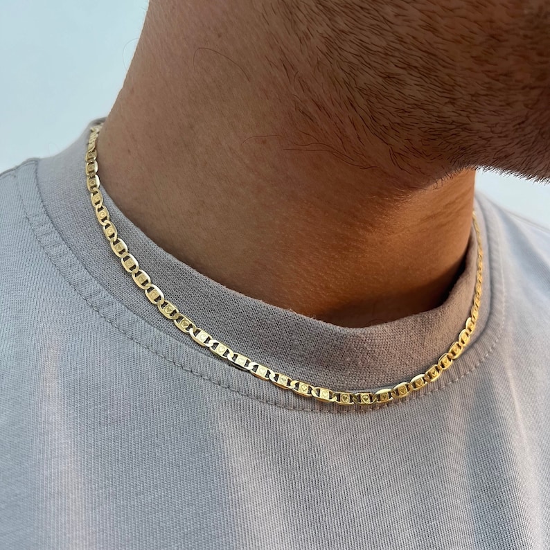 Może przedstawiać: Złoty naszyjnik z łańcuszkiem w stylu marynarskim. Łańcuszek nosi osoba w jasnoszarym t-shircie.