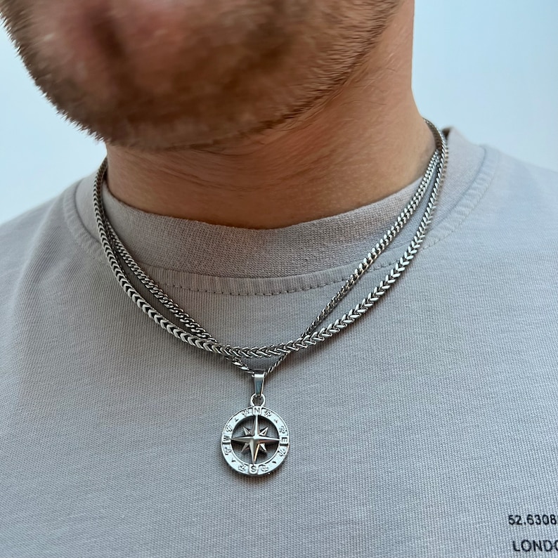 May include: A silver-colored necklace with a compass pendant. The pendant features a star design within a circular frame, suspended from a chain. The necklace is layered over a second, similar chain. The background is a gray shirt.