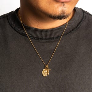 May include: Gold-colored necklace with two pendants. One pendant is an oval shape with a religious figure, and the other is a small cross. The necklace is worn against a dark gray shirt.
