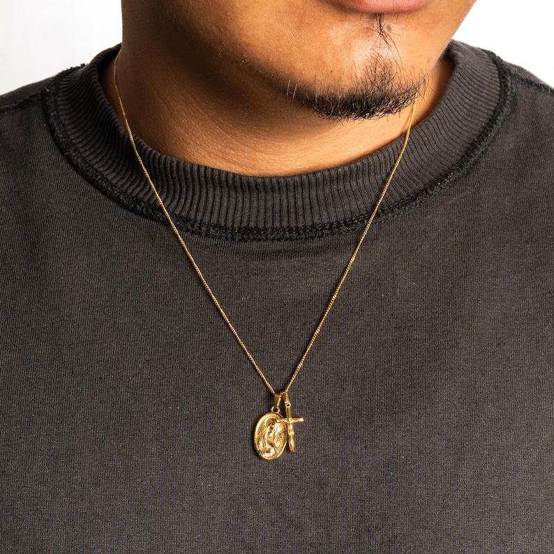 May include: Gold-colored necklace with two pendants. One pendant is an oval shape with a religious figure, and the other is a small cross. The necklace is worn against a dark gray shirt.