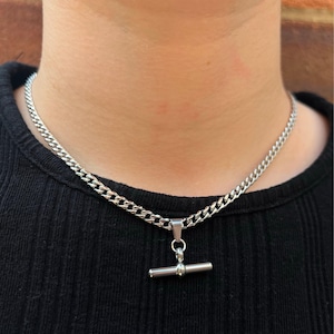 May include: A silver chain necklace with a silver bar pendant. The chain is a curb chain style.