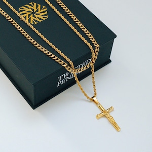 May include: Gold-toned necklace with a cross pendant, displayed on a black box. The necklace features a rope chain and a curb chain. The box has a gold geometric design and the text "TWISTED PENDANTS".