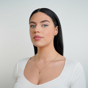 May include: A woman with long black hair wearing a white shirt with a gold chain necklace with a small cross pendant.