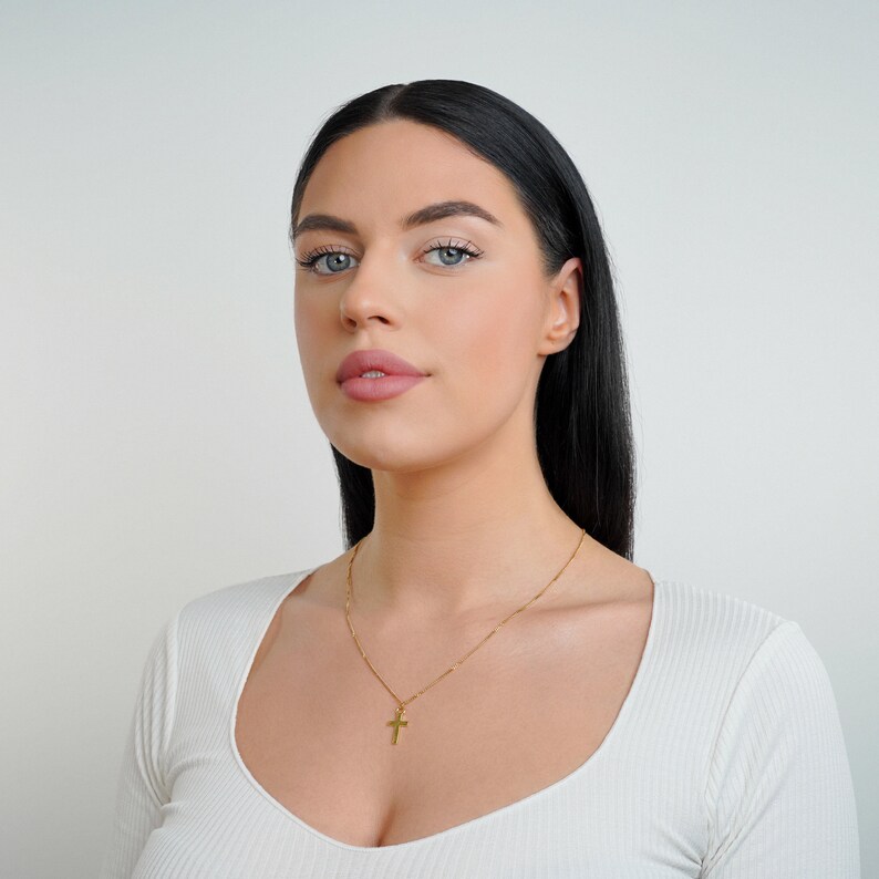 May include: A woman with long black hair wearing a white shirt with a gold chain necklace with a small cross pendant.
