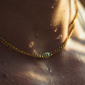 Peut inclure: Un collier chaîne en or avec un pendentif carré en pierre précieuse bleue. Le collier est de style chaîne gourmette, et le pendentif est centré sur la poitrine. Des gouttelettes d'eau sont visibles sur la peau.