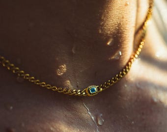 May include: A gold chain necklace with a square blue gemstone pendant. The necklace is a curb chain style, and the pendant is centered on the chest. Water droplets are visible on the skin.