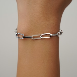 May include: A silver-colored link bracelet with a rectangular chain design. The bracelet is worn on a wrist and has a polished finish. The links are connected in a repeating pattern, creating a modern and minimalist aesthetic.
