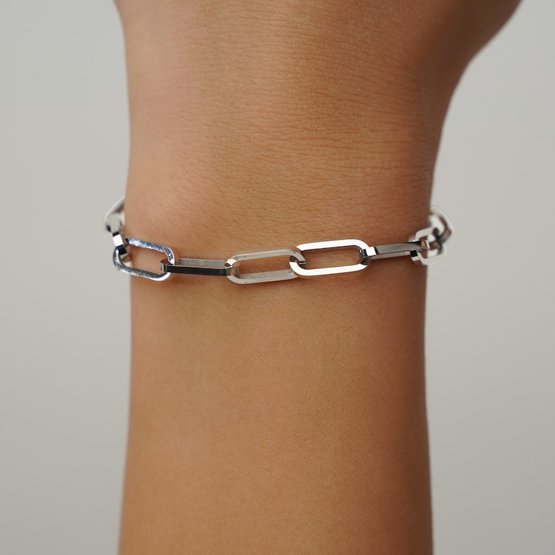May include: A silver-colored link bracelet with a rectangular chain design. The bracelet is worn on a wrist and has a polished finish. The links are connected in a repeating pattern, creating a modern and minimalist aesthetic.
