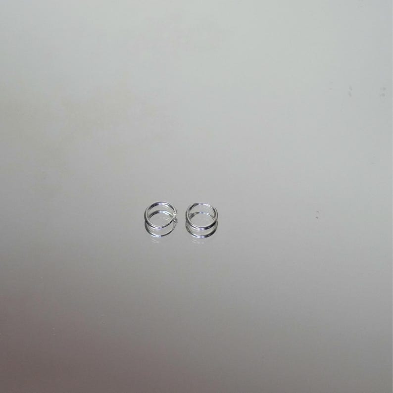 Puede incluir: Dos peque&ntilde;os pendientes de aro plateados. Cada pendiente presenta un dise&ntilde;o de doble banda, creando un efecto de capas. Los pendientes se exhiben sobre una superficie reflectante, realzando su brillo y detalle. El dise&ntilde;o minimalista sugiere simplicidad y elegancia.