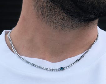 May include: A silver chain necklace with a small, rectangular, teal-colored stone. The necklace is made of interlocking curb links and is worn against a white t-shirt. The necklace is a stylish accessory.