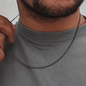 Pode incluir: Um colar de corrente cinza escuro sobre uma camisa cinza. O colar é uma corrente fina e delicada. A imagem é um close-up do colar e da pessoa que o usa.