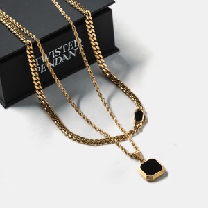 May include: Gold-toned layered necklaces with a black square pendant. The necklaces have different chain styles, including a rope chain and a curb chain. The pendant is a black square with a gold border. The necklaces are displayed on a white surface with a black box in the background.