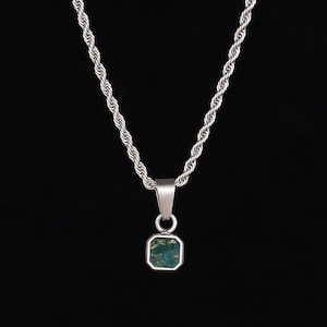 May include: Silver-toned necklace with a rope chain and a square pendant. The pendant features a green and blue stone set in a silver frame. The necklace hangs against a black background, highlighting the jewelry's design.