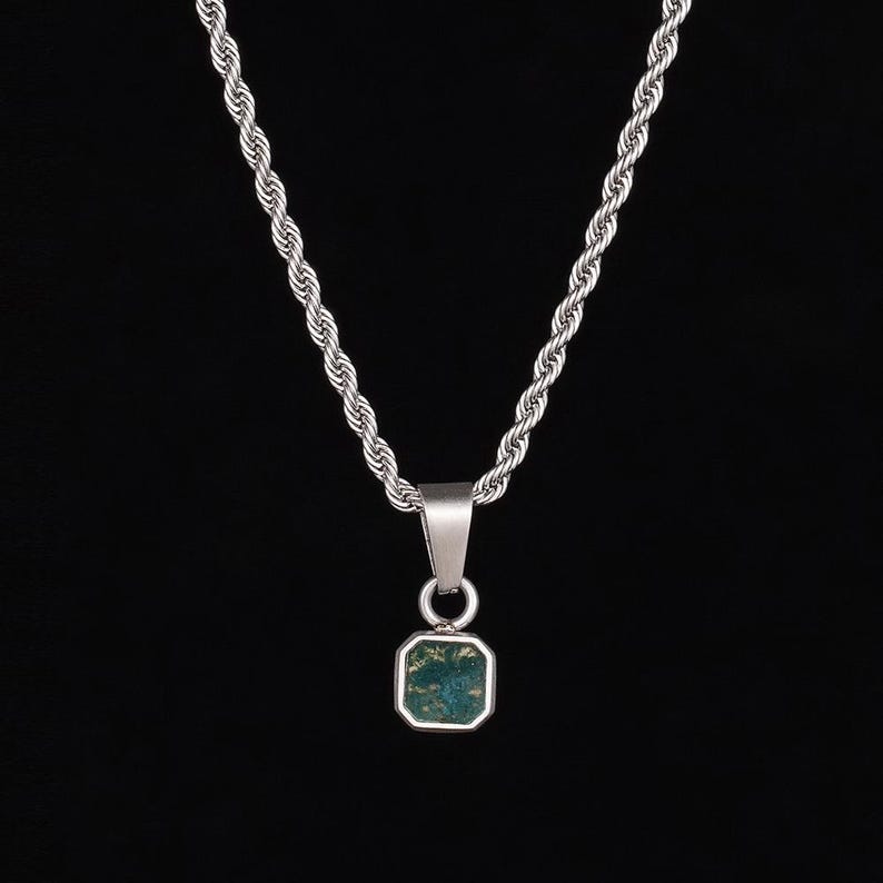 May include: Silver-toned necklace with a rope chain and a square pendant. The pendant features a green and blue stone set in a silver frame. The necklace hangs against a black background, highlighting the jewelry's design.