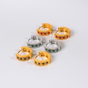 May include: Six hoop earrings in gold and silver tones. The gold earrings feature black star designs, while the silver earrings have green star designs. The other gold earrings have red star designs. All earrings have a polished finish.
