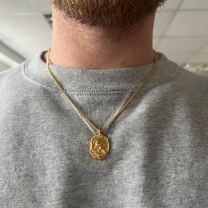 May include: A gold chain necklace with a round pendant featuring a profile of a person wearing a helmet. The pendant is about 1 inch in diameter.