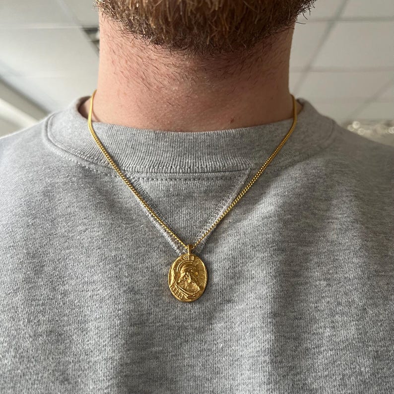 May include: A gold chain necklace with a round pendant featuring a profile of a person wearing a helmet. The pendant is about 1 inch in diameter.