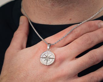 May include: A silver-toned circular pendant featuring a compass rose design with embedded clear stones, suspended from a matching silver-toned rope chain necklace. The pendant has a polished metallic finish and a detailed star-shaped centre.