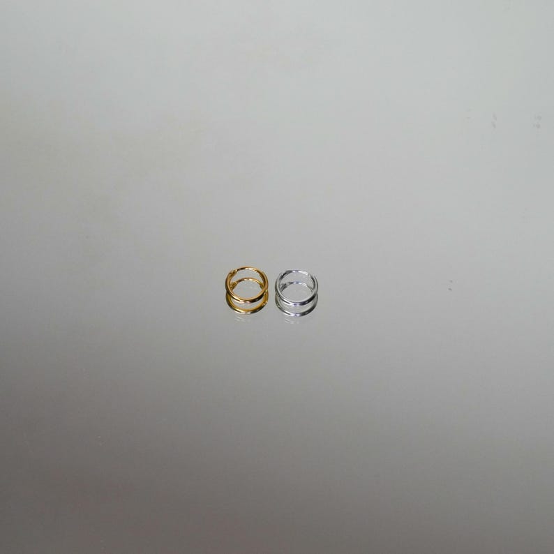 Puede incluir: Se muestran dos anillos, uno dorado y otro plateado. Cada anillo presenta un dise&ntilde;o de doble banda. Los anillos se colocan sobre un fondo reflectante y neutro, destacando su elegante sencillez. Estos anillos son adecuados para el uso diario.