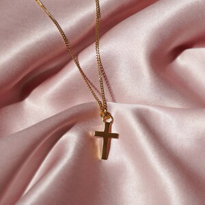 May include: A gold chain necklace with a simple gold cross pendant. The necklace is laying on a pink satin fabric.