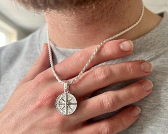 May include: A silver-toned rope chain necklace featuring a circular pendant with a compass rose design. The pendant has a textured border and a central star-like motif with four cardinal directions clearly engraved on its metallic surface.