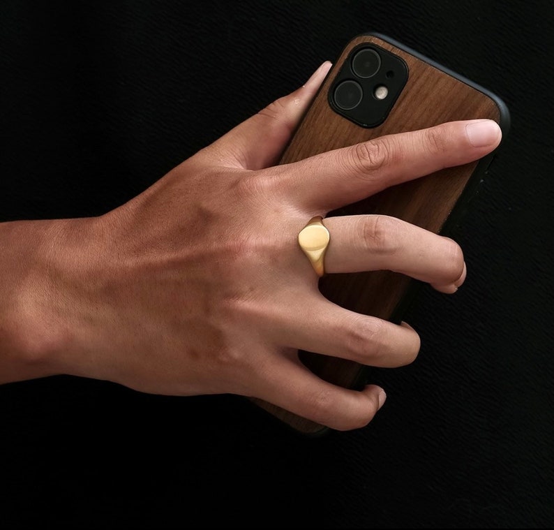 May include: A gold signet ring with a smooth oval top. The ring is worn on the index finger of a hand holding a brown leather phone case.