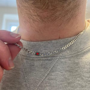 Puede incluir: Un collar de cadena de plata con un colgante rectangular de piedra roja. El collar se muestra sobre un fondo gris, mostrando los eslabones de la cadena y el color del colgante.