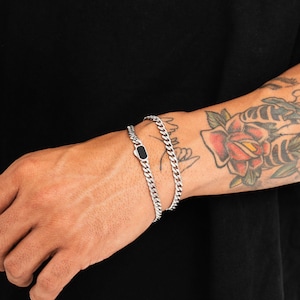 Könnte beinhalten: Ein silberfarbenes Panzerkettenarmband mit einem einzelnen rechteckigen schwarzen Emaille-Inlay-Detail. Das Armband besteht aus ineinandergreifenden Metallgliedern, die eine durchgehende Kette bilden und eine polierte, reflektierende Oberfläche aufweisen.