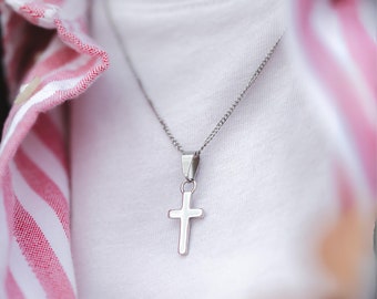 May include: A silver cross pendant necklace. The cross is a simple, polished design, suspended from a delicate chain. The necklace is worn over a white shirt, with a red and white striped shirt collar visible in the background.